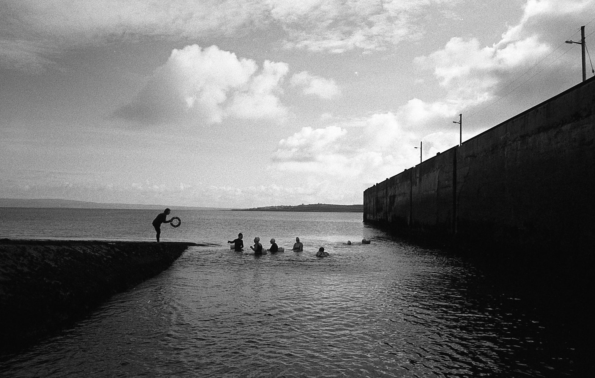 Black and white photo of a person jumping into water from a concrete wall with people waiting below.