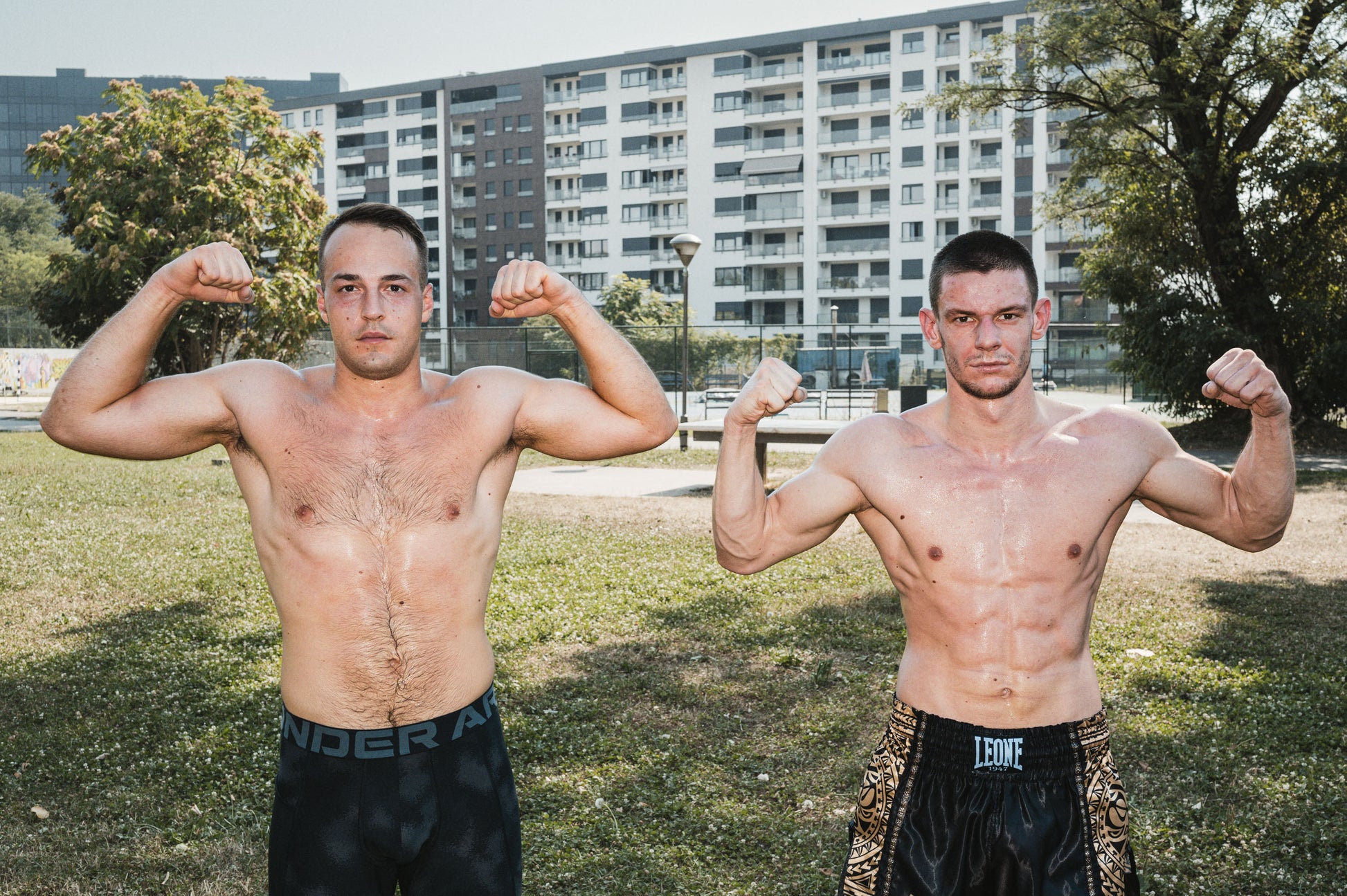 Two men flexing their muscles outdoors with a building and trees in the background